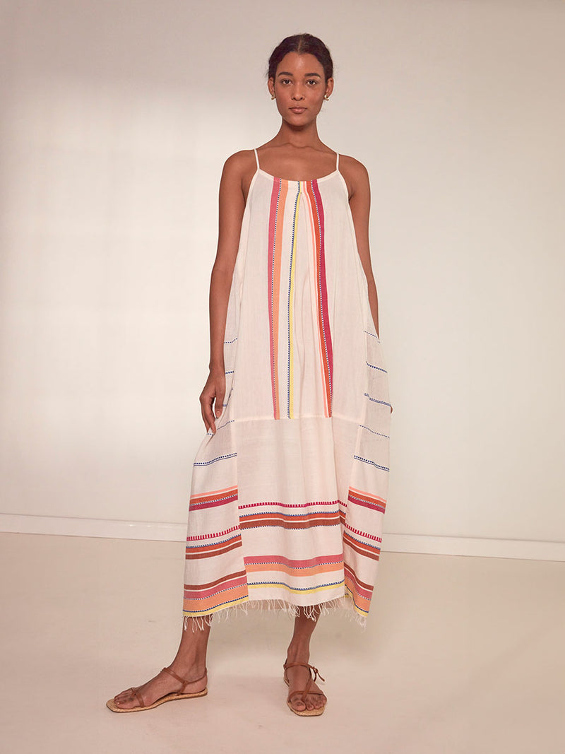 Woman Standing Wearing  lemlem Nia Slip Dress featuring tibeb inspired stripes in a vibrant fiesta of colors against a creamy vanilla background.