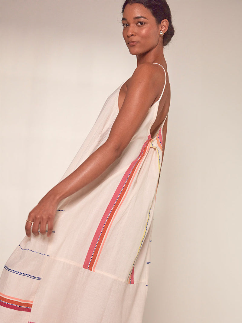 Side View of a Woman Standing Wearing lemlem Nia Slip Dress featuring tibeb inspired stripes in a vibrant fiesta of colors against a creamy vanilla background.