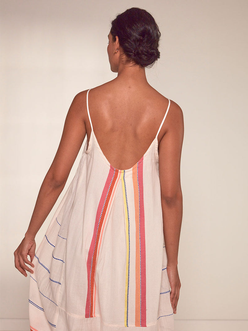 Back View of a Woman Standing Wearing lemlem Nia Slip Dress featuring tibeb inspired stripes in a vibrant fiesta of colors against a creamy vanilla background.