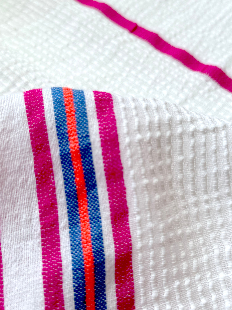 Close up on zelele fabric featuring stripe pattern of bright magenta, blue, and orange on s crispy white background. 