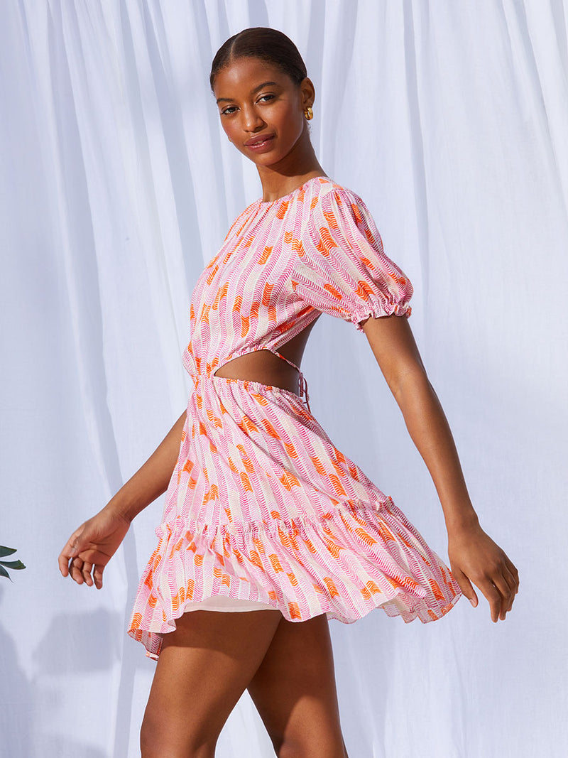 A side view of a woman walking the Gigi Short Open Back Dress in Multi Pink featuring pink, peach and orange allover chevron print and strips of gold lurex.